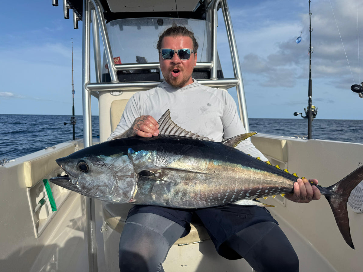 Sportfiskare med solglasögon och vit skjorta håller en stor tonfisk på båt. Havet och fiskeutrustning syns i bakgrunden.