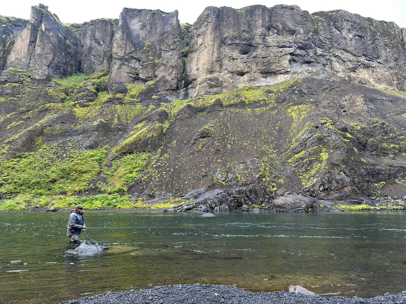 Sportfiskare fiskar i lugn älv, höga mossklädda klippor i bakgrunden och stenig strand-perfekt för en unik fiskeresa.