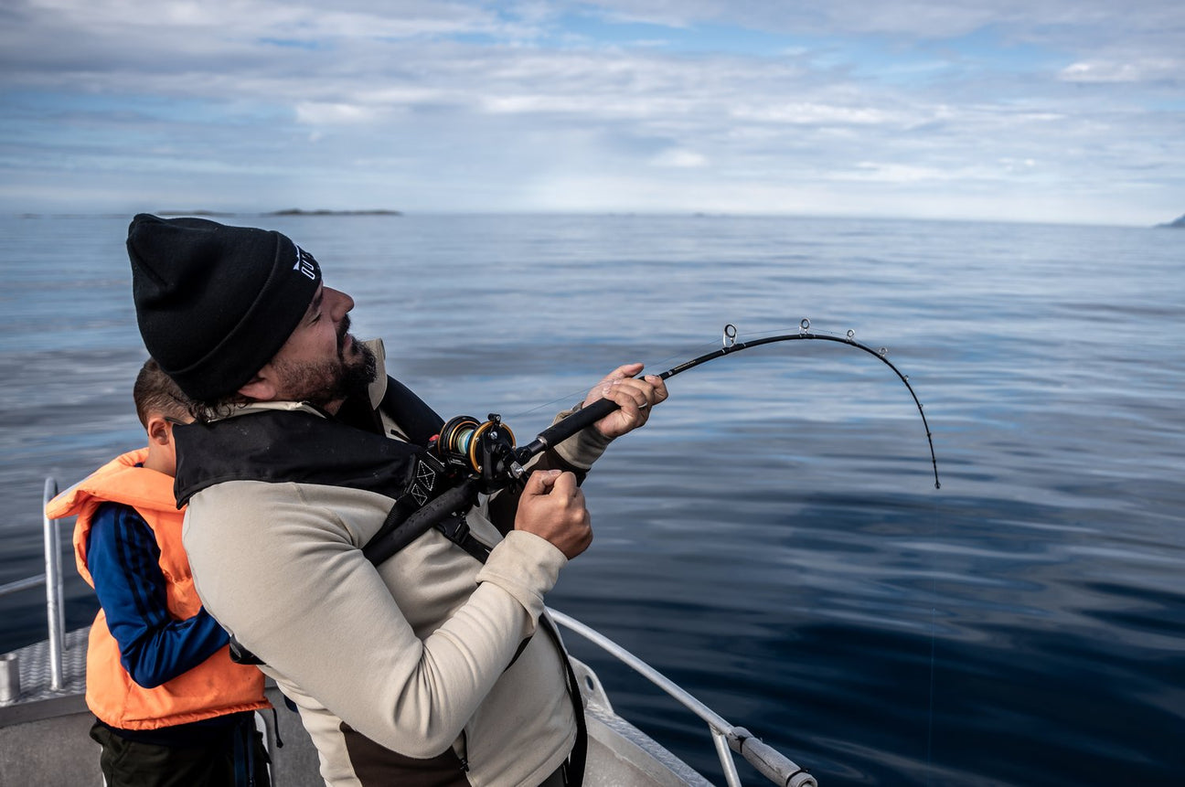 En man med mössa och flytväst står på en båt och håller i ett spänt fiskespö, medan ett barn i orange flytväst står bredvid honom över lugnt, öppet vatten.