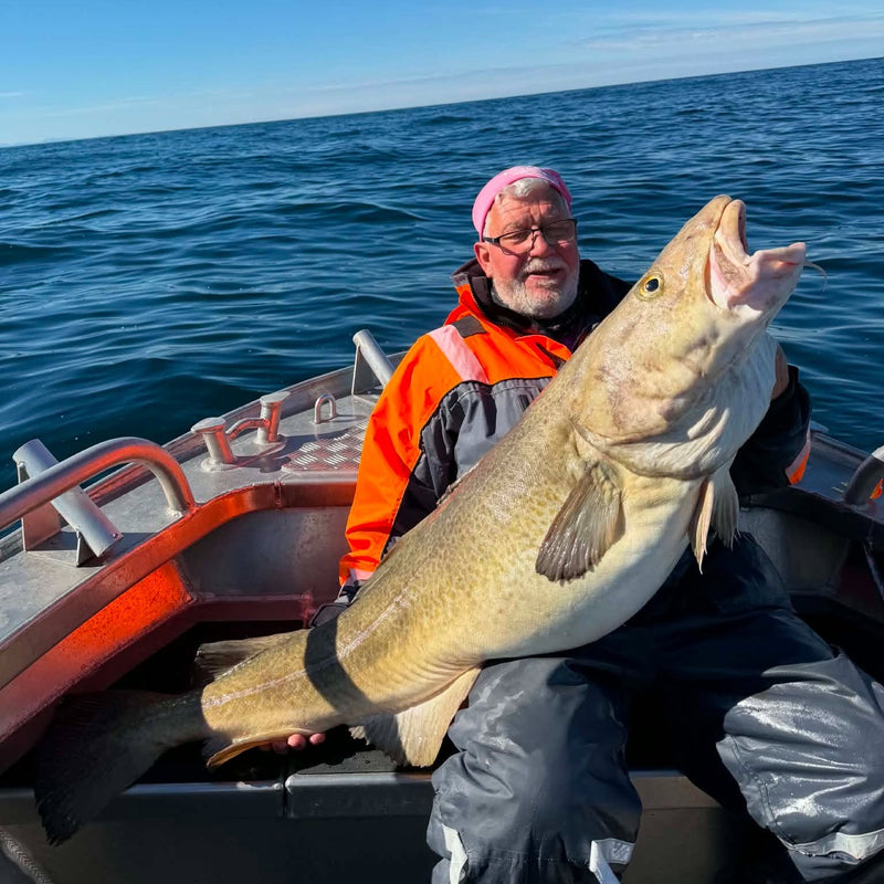 En person i båten håller upp en stor fisk och ler mot kameran under sin fisketur. Klar himmel och lugnt hav skapar perfekta förutsättningar för fiske.