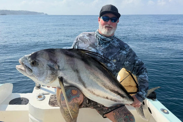 En sportfiskare med hatt och solglasögon håller upp en stor silverfisk under fiskeresa, med havet i bakgrunden.