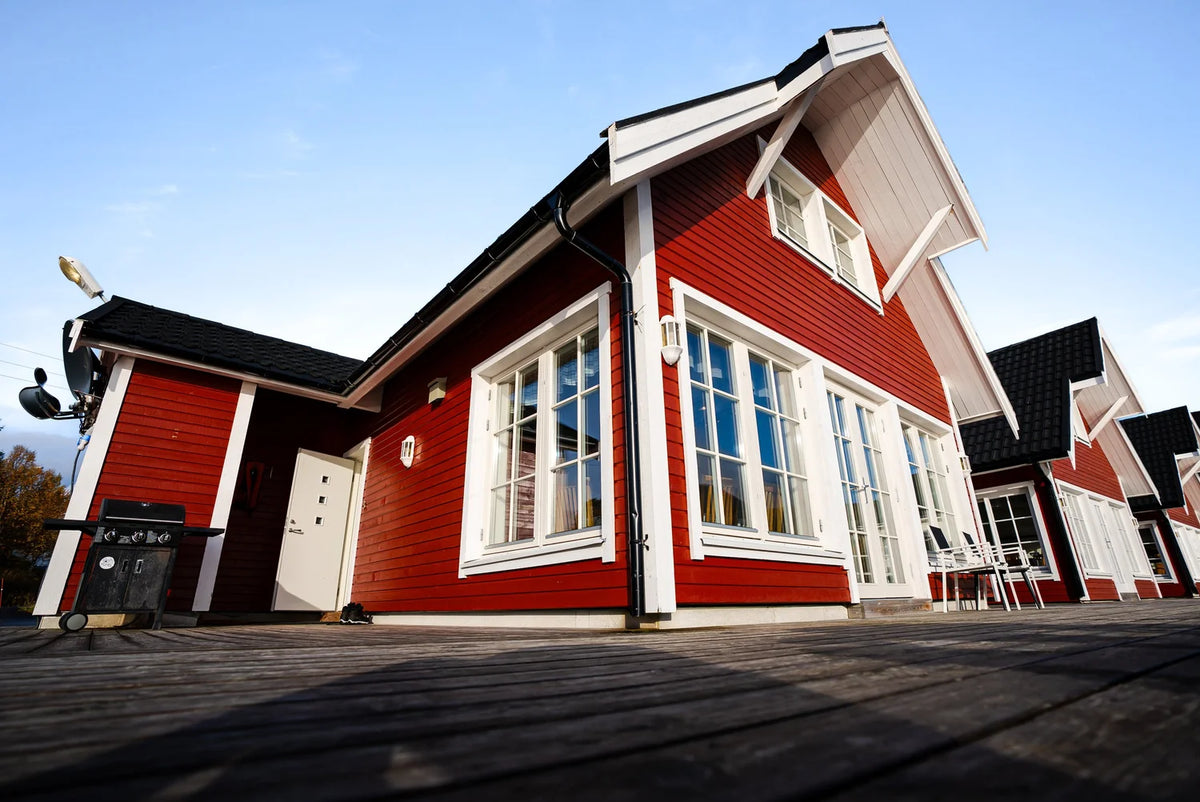 Rött trähus med vita knutar, stora fönster och svart tak på trädäck under blå himmel-idealisk vila för sportfiskare.