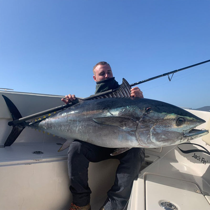 En sportfiskare sitter på en båt och håller stolt upp en stor tonfisk mot klarblå himmel, perfekt för fiskeäventyr.