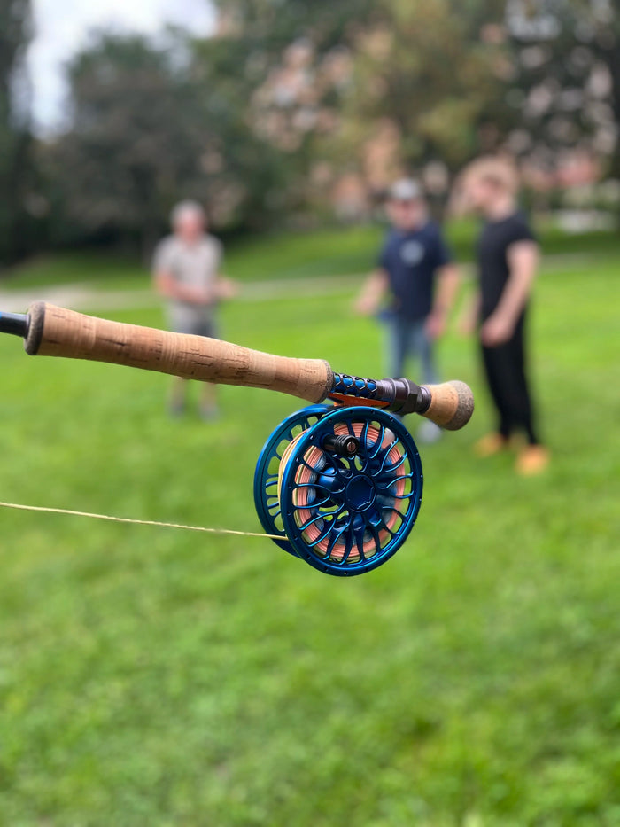 Närbild på fiskespö och rulle, tre suddiga personer på gräs i bakgrunden - sportfiske och fiskeresa i fokus.