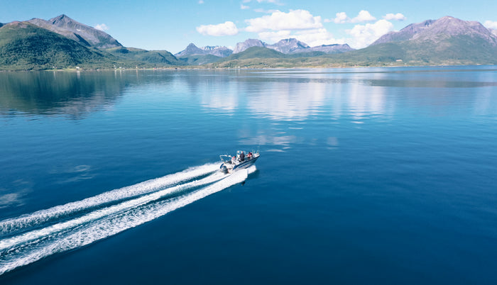 En liten motorbåt kör snabbt över en stilla blå sjö med vitt svall. Gröna berg och blå himmel speglar sig-idealisk fiskeresa.