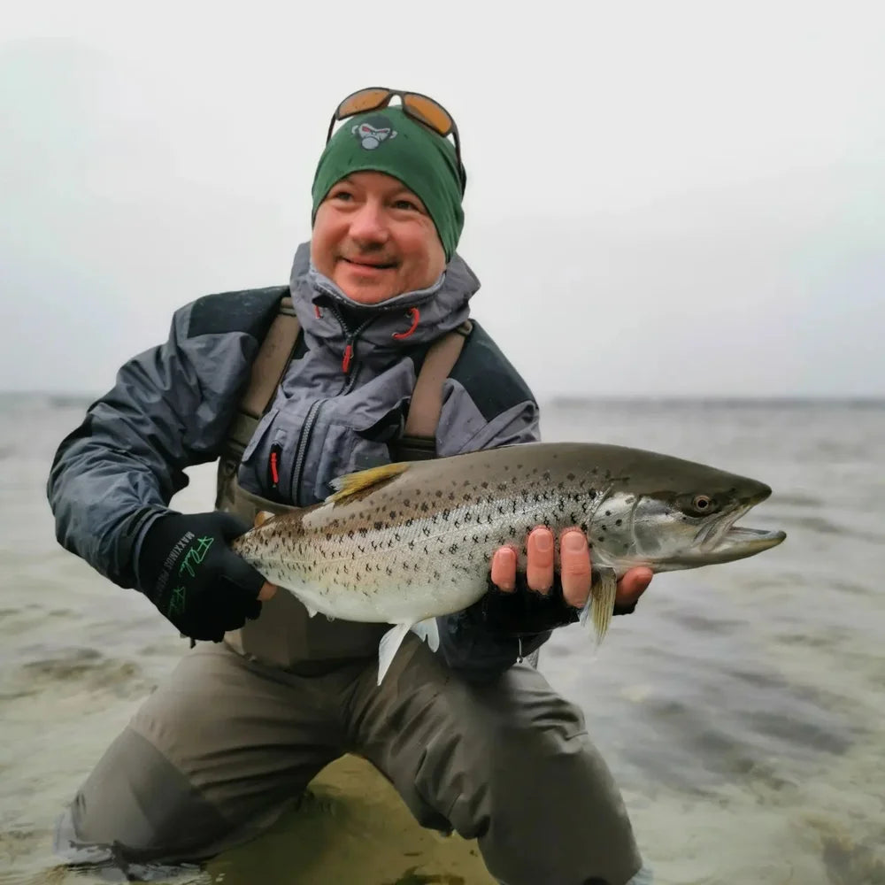 En sportfiskare i outdoor-kläder knäböjer i grunt vatten, ler och håller en stor prickig fisk. Grå himmel ger äventyrskänsla.