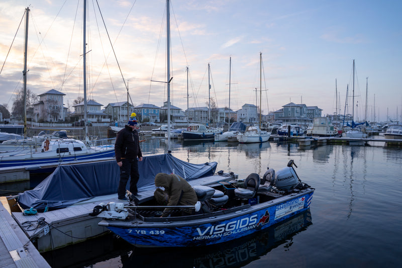 Två sportfiskare står på en liten blå fiskebåt vid bryggan, omgivna av segelbåtar och sjönära hus i gryning eller skymning.