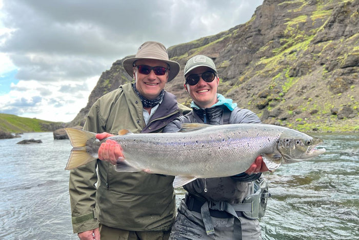 Två sportfiskare i outdoor-kläder håller gemensamt upp en stor fisk vid en älv omgiven av klippor och grönska på sin fiskeresa.