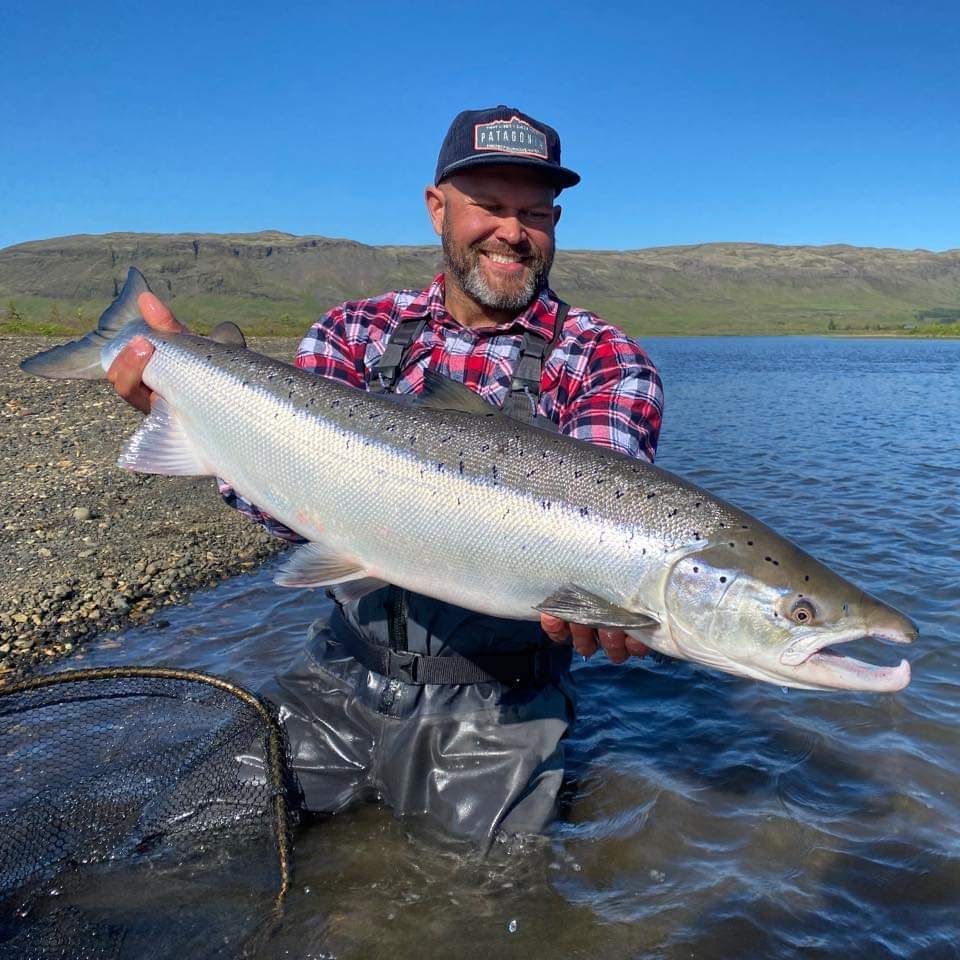 Leende sportfiskare i vadarbyxor och rutig skjorta håller en stor silverfisk vid en älv, med håv och gröna kullar i bakgrunden.