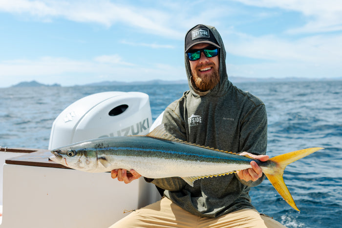 Man med solglasögon och hoodie ler och håller en stor yellowtail-fisk ombord på båt under fiskeresa, hav och blå himmel i bakgrunden.