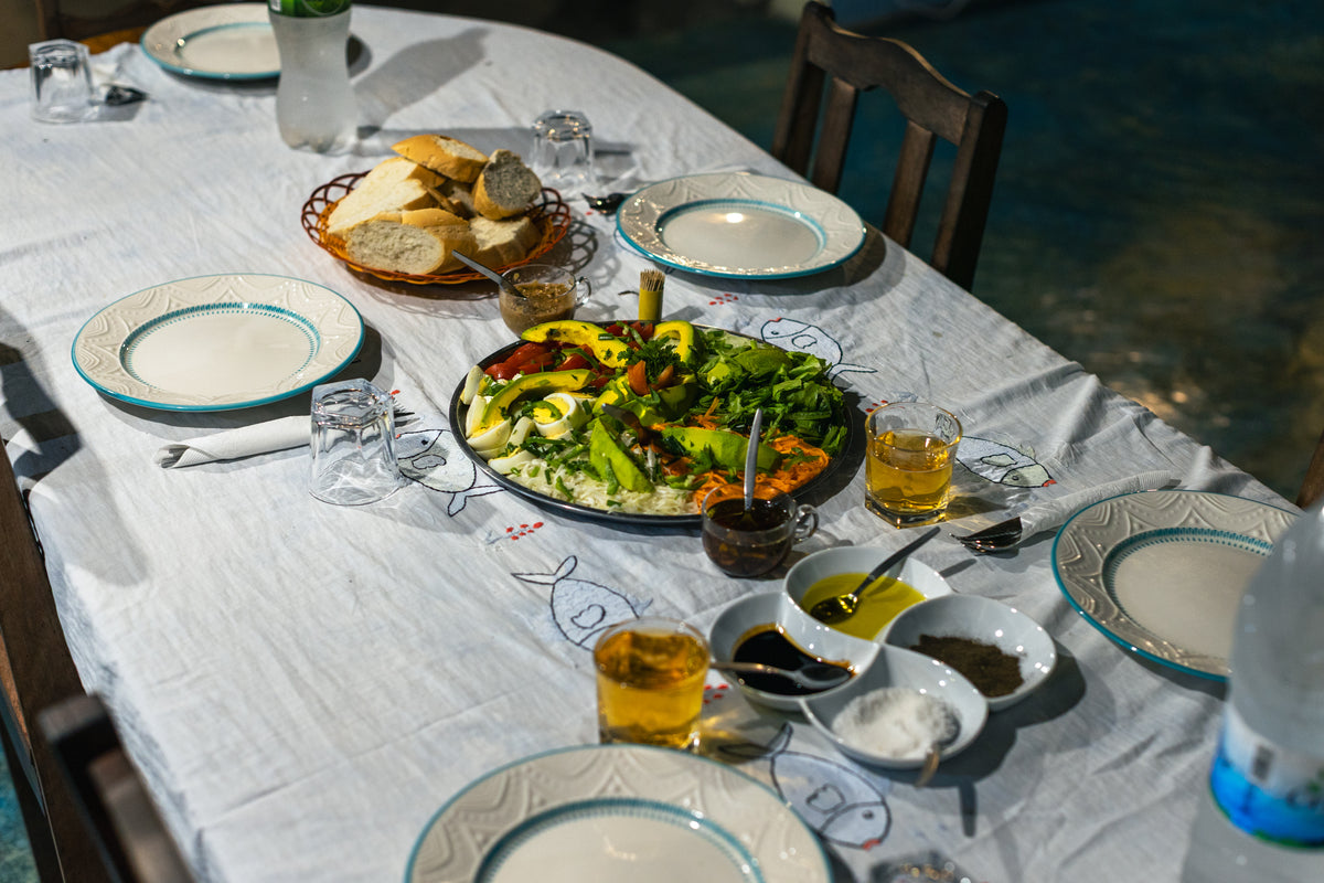 Dukat matbord med tallrikar, juiceglas, bröd och sallad på vit duk med fiskmotiv - perfekt för sportfiske- och resesällskap.
