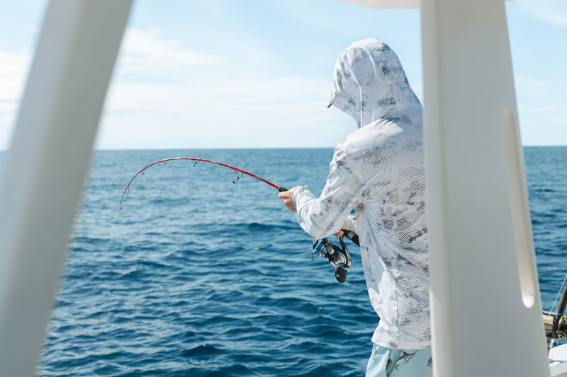 En sportfiskare i huvtröja fiskar från båt på en fiskeresa, håller ett böjt spö över blått hav under delvis molnig himmel.