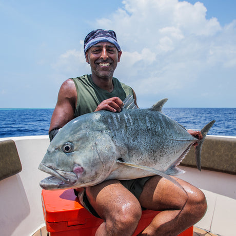 Sportfiskare med stor fisk i knät på båt, omgiven av hav och moln under en fiskeresa Maldiverna Popping  Jigging.