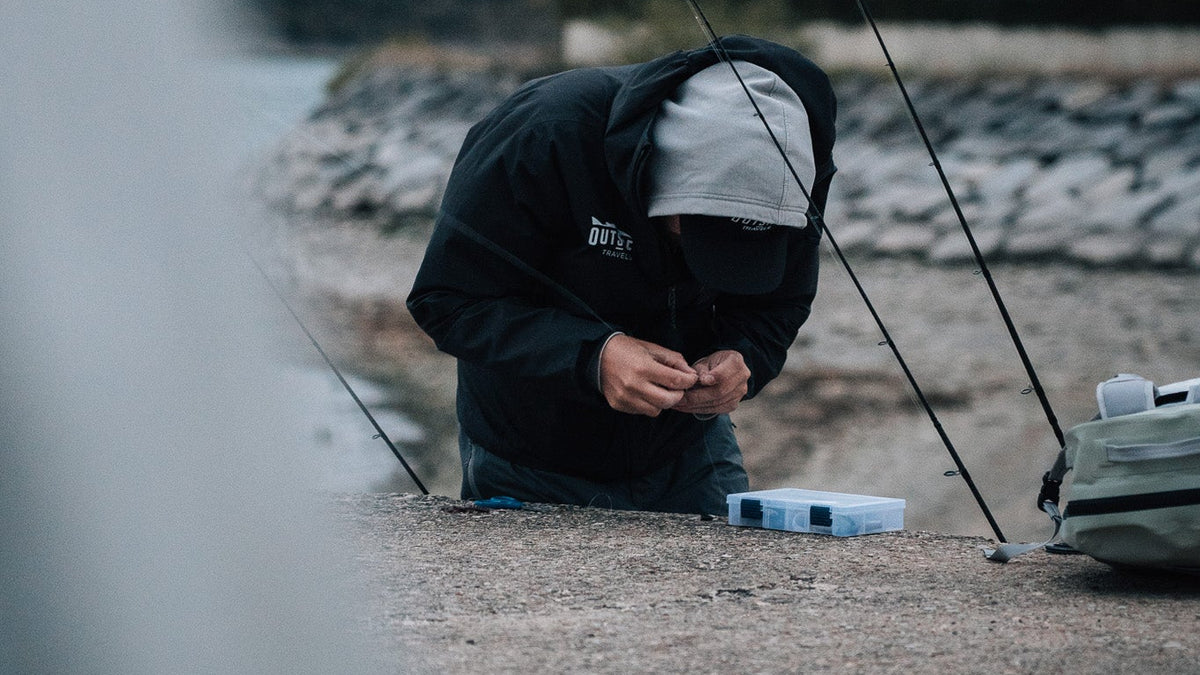 En sportfiskare i huva och keps förbereder fiskeutrustning vid vattnet, med beteslåda och fiskespön under sin fiskeresa.