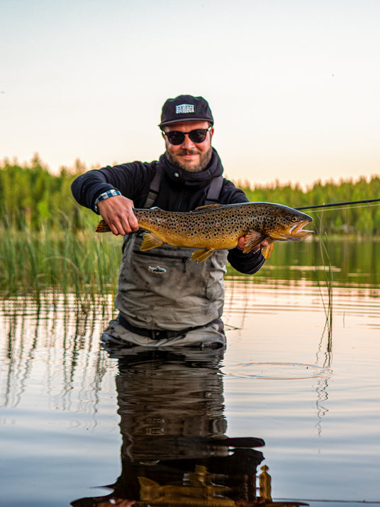 Leende sportfiskare i vadarbyxor håller en stor brunöring, omgiven av lugn sjö och skog-en höjdpunkt på fiskeresan.