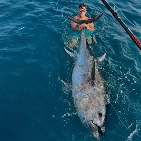 Sportfiskare i badbyxor fiskar med spö bredvid stor tonfisk i klart vatten på Fiskeresa Ebrodeltat, Spanien.