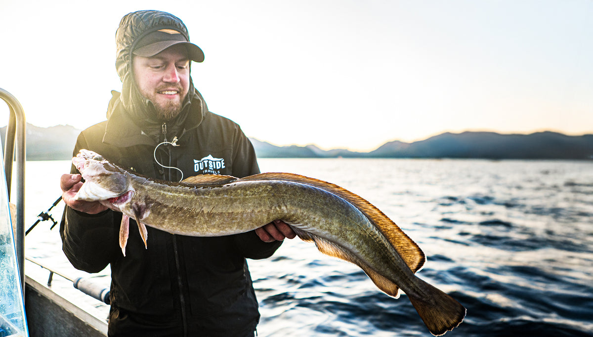 En sportfiskare på båt håller en stor fisk och ler mot kameran. Solnedgång över berg och lugnt vatten i bakgrunden.